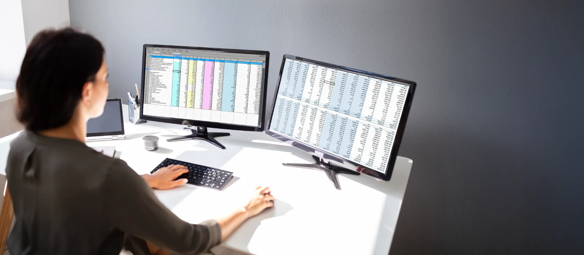Woman working at desk with dual monitors displaying spreadsheets and business data for financial products subledger