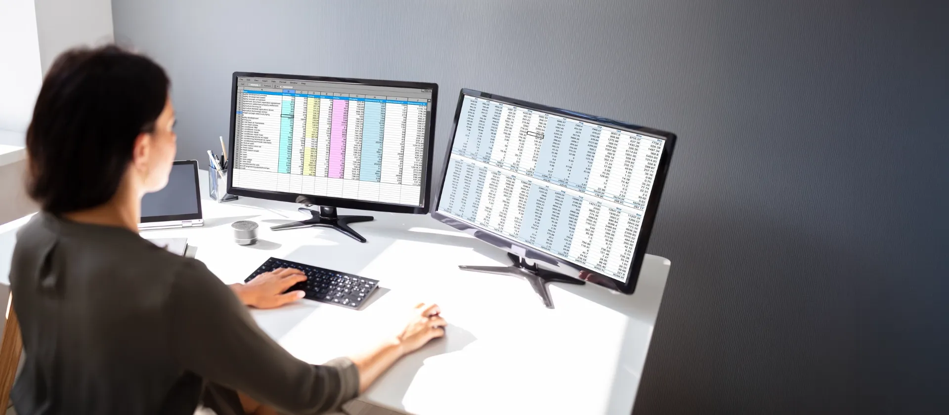 Woman working at desk with dual monitors displaying spreadsheets and business data for financial products subledger