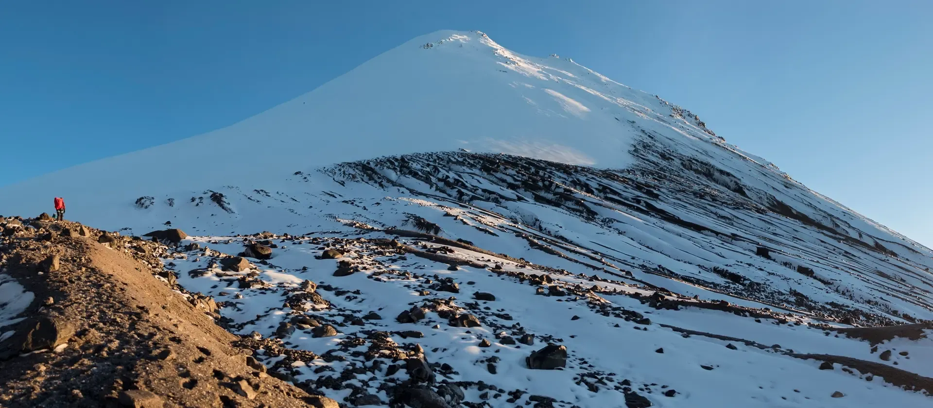 Hiker in red jacket climbing snowy mountain slope toward a sunlit peak under a clear blue sky