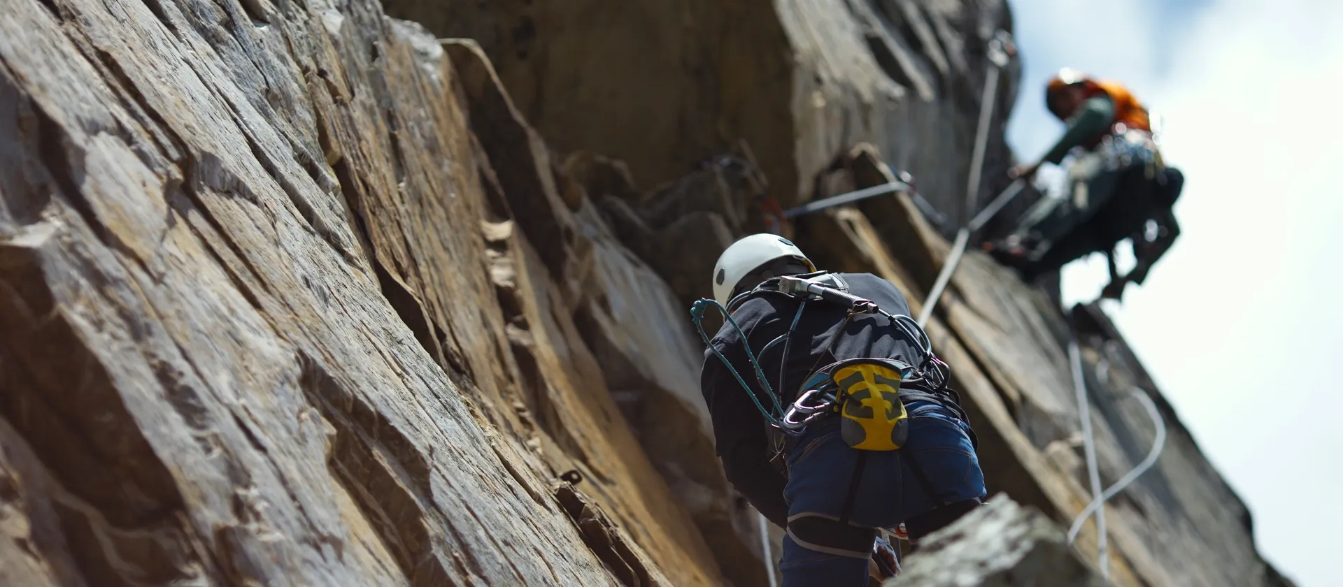 Two climbers ascending a steep rock face with helmets and safety ropes during an outdoor adventure.