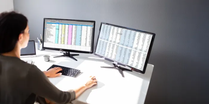 Woman working at desk with dual monitors displaying spreadsheets and business data for financial products subledger